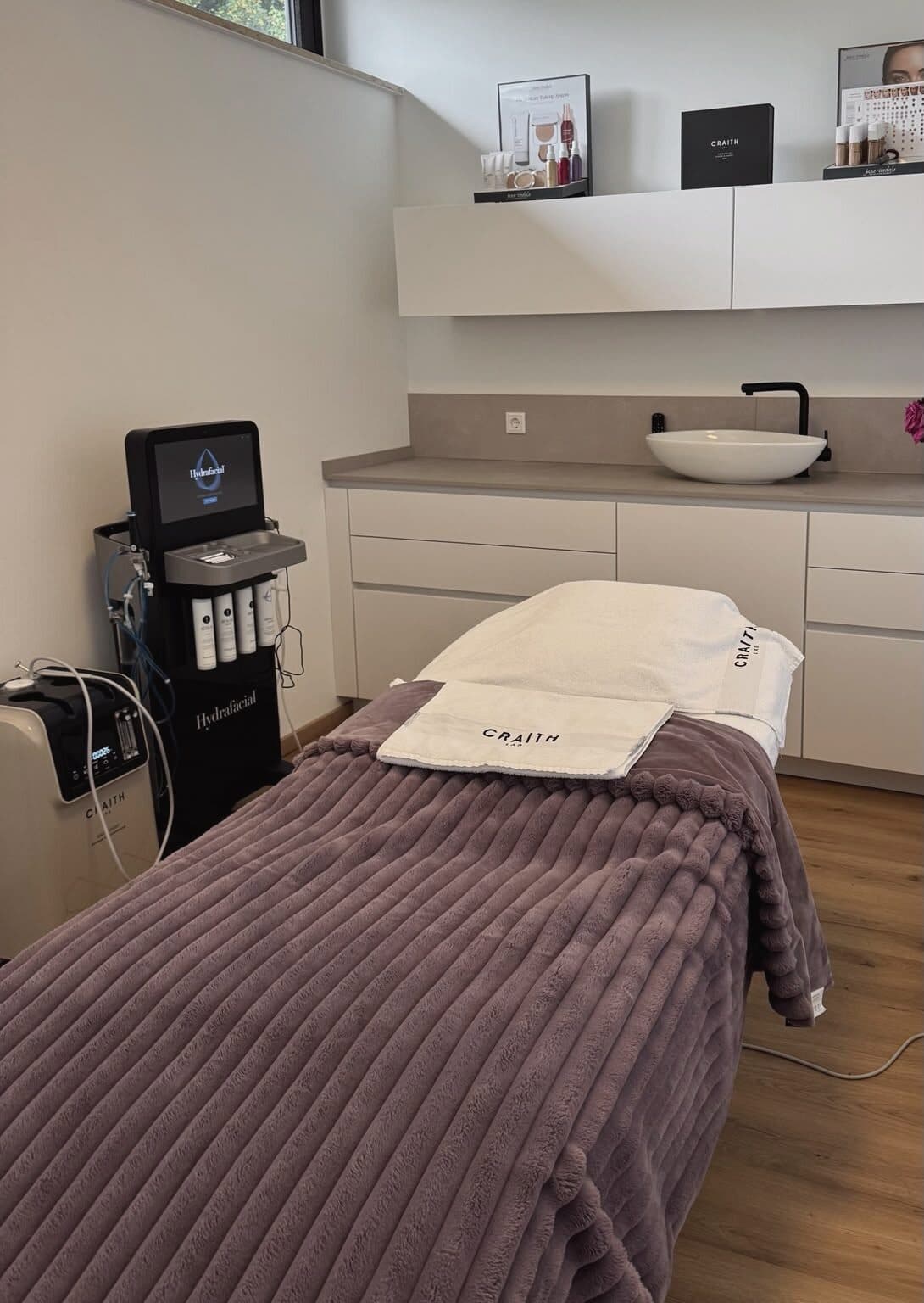 Behandlungsraum im Beautyloft: Liege mit gerippter Decke in Mauve, HydraFacial-Gerät, Waschtisch und Pflegeprodukte im Regal