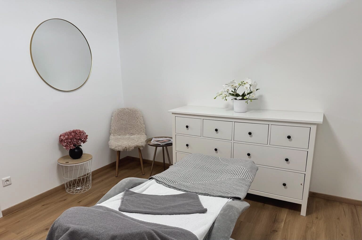 Heller Behandlungsraum im Beautyloft mit Liege, Kommode, weissen Blumen und rundem Spiegel