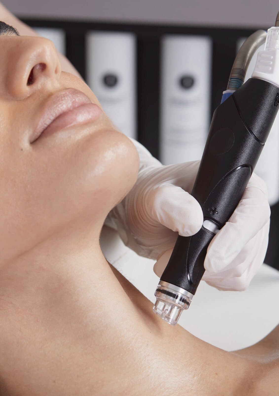 Nahaufnahme: Handstück eines HydraFacial-Geräts am Hals unter dem Kinn, im Hintergrund Serumflaschen