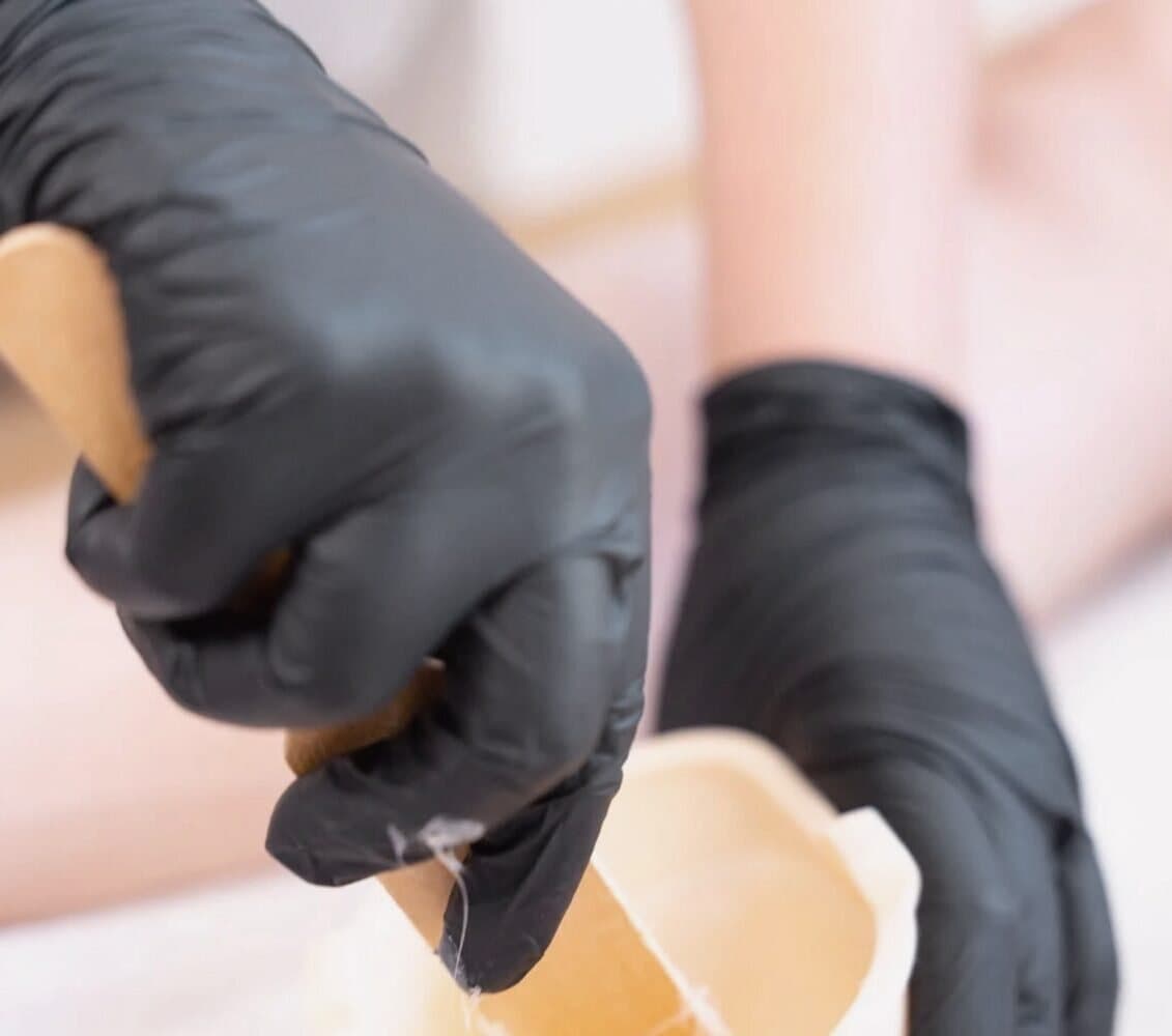 Hände in schwarzen Handschuhen nehmen mit einem Holzspatel Wachs aus dem Topf