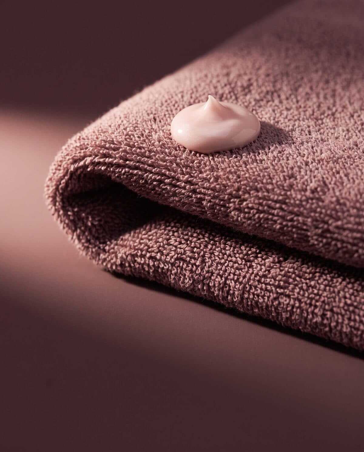 Gefaltetes Frotteetuch in Mauve, darauf ein Tupfer rosafarbener Creme, seitlich einfallendes Licht